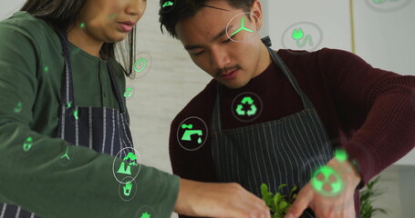 Arranging green seedlings in planter by Asian team in aprons at studio countertop with eco icons