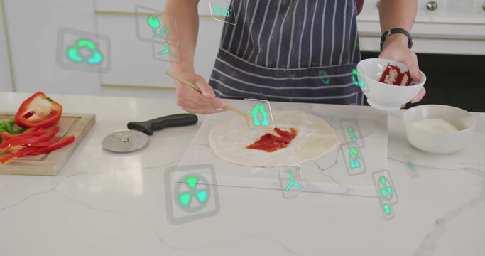 Teenage boy wearing apron smearing tomato sauce on dough in home kitchen, with spoon, eco icons - Powered by Adobe