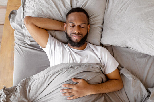 Healthy Sleeping Concept. Above top view portrait of happy young well-slept African American man lying on back in bed with closed eyes, resting in bedroom in the morning, having hap