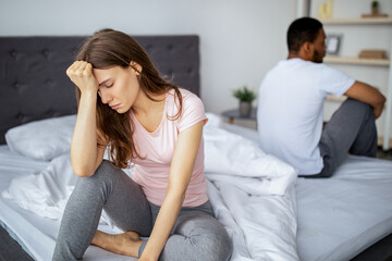Interracial married couple having family difficulties, sitting on opposite sides of bed, feeling...