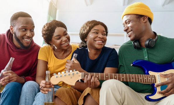 Cute black friends playing guitar and singing songs while having party at home, sitting on couch in living room, drinking beer, joyful african american people two couples relaxing at weekend - Powered by Adobe