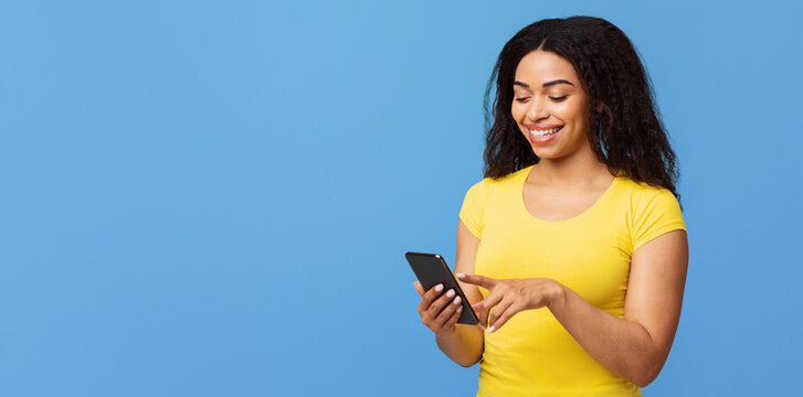 Cool app. Happy african american woman using smartphone, texting or scrolling social networks feed, standing over blue studio background with free space
