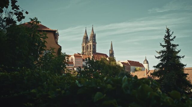 View of Albrechtsburg Castle and Meissen Cathedral rising above the old town rooftops, framed by greenery under a clear summer sky in Saxony, Germany.