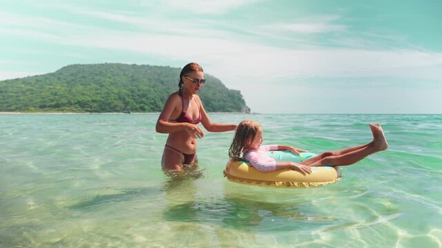 Mom and daughter laugh together as girl floats in donut ring. Pure beach joy, captured in turquoise water under sunny sky. Ideal image of summer vacation and loving family moment.