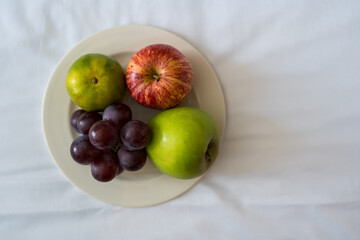 A plate of grapes, an orange,a and a red and green apple on white plate sitting on a bed at a resort