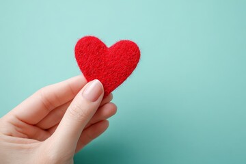 Fototapeta premium A tender red heart-shaped felt object is gently held by a hand against a tranquil teal backdrop.