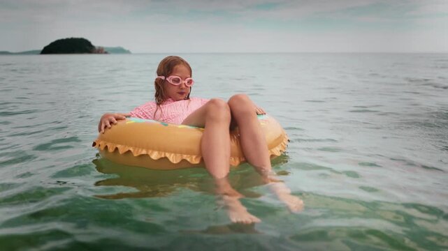 Young girl child in goggles floating on colorful inflatable donut in crystal-clear sea water. Joyful summer moment of carefree childhood during beach holiday.