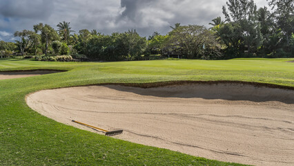 Golf course. Green trimmed grass on the field. Rake on clean sand in a hollow. Tropical vegetation in the distance. Clouds in the sky. Mauritius. Hotel. Resort.