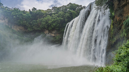 Fototapeta premium Beautiful tropical waterfall. Streams of water flow down from a sheer cliff into the river. Foam, splashes, fog. Green vegetation on the rock. Clouds in the sky. China. Huangguoshu Waterfall. Guizhou