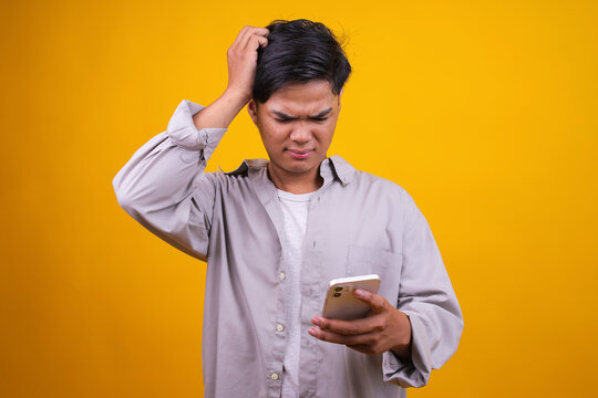 Asian Man Holding Smartphone in Hand With Confused Expression Against Yellow Background