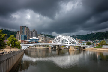 White arched bridge spans a calm river under dramatic stormy clouds, with urban buildings and green hills in the background, reflecting a moody cityscape scene