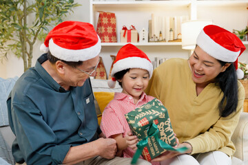 Asian grandparents giving Christmas gift to grandson smiling happily sitting together wearing santa hats joyful festive family holiday cozy warm home moment, Concept of happiness festival