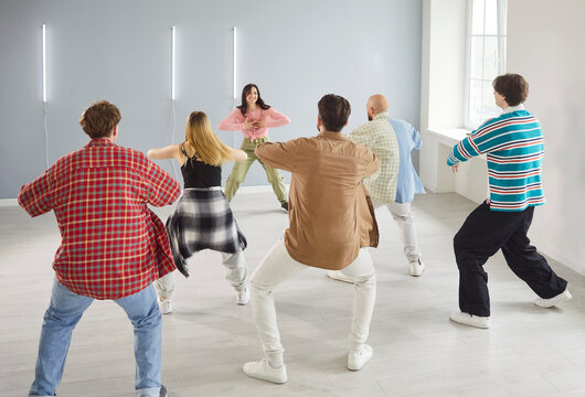 Modern indoor dance studio class, diverse group of dancing young people actively following trainer woman learning dance steps, team moving practicing choreography, feeling energized, rear view 