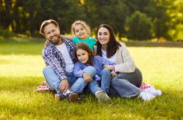 Fototapeta premium Portrait of happy smiling family with son and daughter sitting on green grass in the summer park and looking cheerful at camera. Mother, father with two kids enjoying sunny day together.