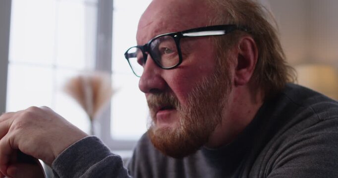 Sad senior man with glasses and beard sitting alone by window at home. Serious lonely old grandfather with unhappy face, stress and anxiety expression, lost elderly man in need of help and care