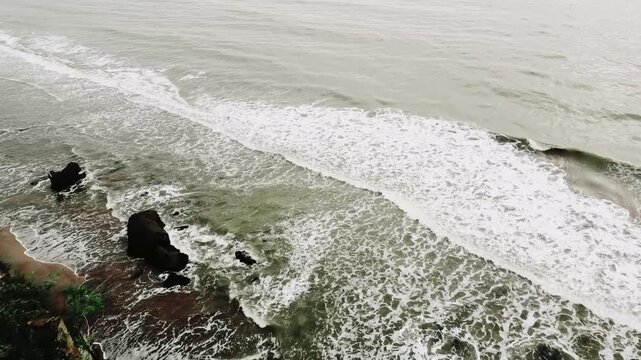 High angle drone shot of powerful ocean waves crashing on a dark rocky beach in Indonesia. A moody, natural seascape with white foam and surf.