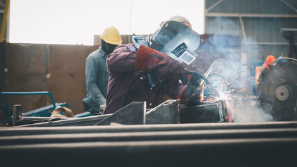 Two skilled workers are engaged in metal welding inside a workshop. One worker uses a welding torch, creating bright sparks and smoke, highlighting the craftsmanship of metal fabrication