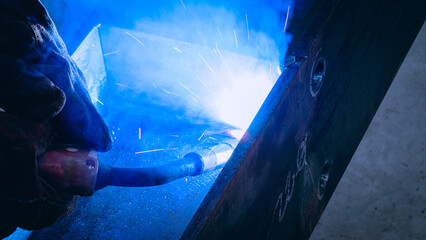 A welder focuses intently on joining metal pieces, bright sparks flying as the welding torch ignites. The workshop setting is dimly lit, enhancing the vibrant light display from the process © Happy Photo
