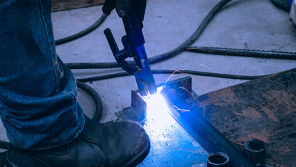 A skilled worker engages in welding, creating sparks that light up the workshop floor. The surrounding tools and materials reflect an industrial atmosphere during the daytime