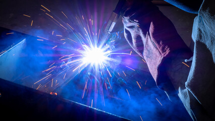 Bright sparks burst in various directions as a welder expertly works on metal, surrounded by a dark...