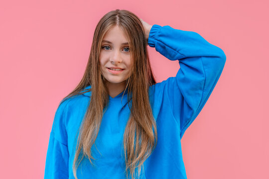 Young girl in hoodie rubs the back of her head with a guilty look, apologetic look, isolated pink background