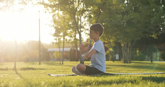Child boy practices yoga and meditation on green lawn in park at sunset. Focused kid in lotus pose with deep breathing promotes healthy lifestyle, fitness, concentration, and mindfulness outdoors.