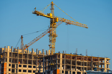 Construction cranes building a modern apartment residential building under construction against a clear blue sky during a sunny day, representing urban development and growth