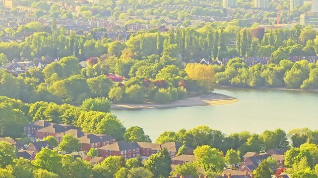 Aerial video of Edgbaston Reservoir in Birmingham, England, with tall summer trees glowing in warm golden light, calm water, and residential suburban homes.