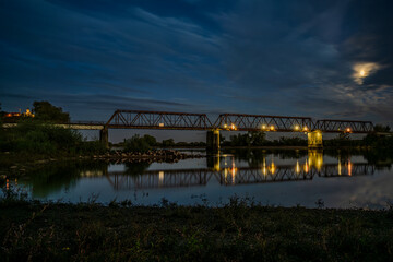 Nachtaufnahme einer Br&uuml;cke &uuml;ber einen ruhigen Fluss mit Lichtreflektionen und sternenklarem Himmel