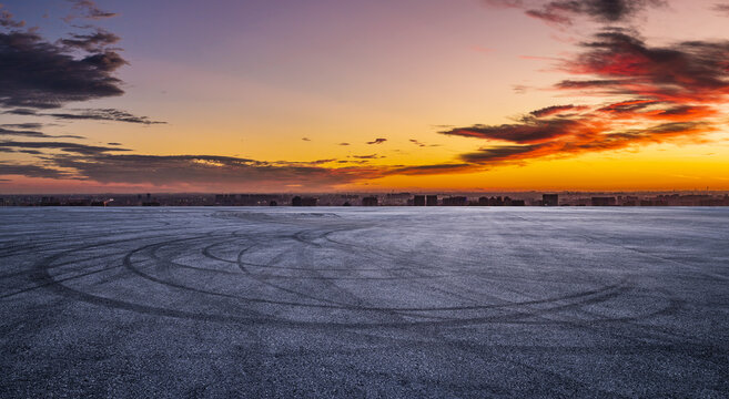 Empty asphalt race track ground with car tire skid marks and city skyline at dramatic sunset