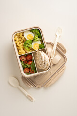 A vertical prepared meal consisting of broccoli, boiled eggs, pasta, rice cakes, and tomatoes, presented in a reusable divided container for convenient and nutritious on-the-go dining