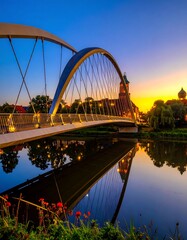 Obraz premium A modern steel arch bridge spans a calm river at sunset, its reflection mirroring the city skyline and bridge structure.
