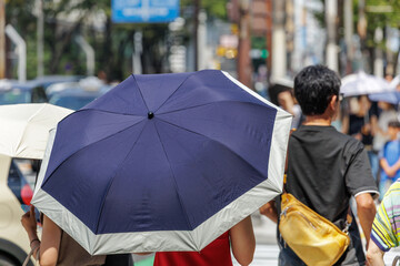 Large umbrella shields person walking along bustling city sidewalk filled with people out enjoying weekend shopping. Scene captured on brilliantly sunny day when urban environment is most lively.