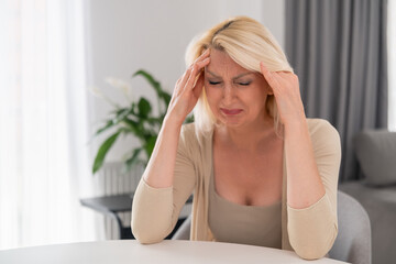 Blonde middle aged woman sitting alone at kitchen table, covering face with hands, crying with...