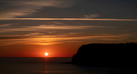 Bright orange sun setting over calm ocean water and dark cliff sunset