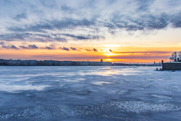 Dramatic winter sunset over a frozen river with colorful sky, clouds, and silhouettes of historical buildings on the horizon. The frozen surface reflects the light of the evening sun, creating a