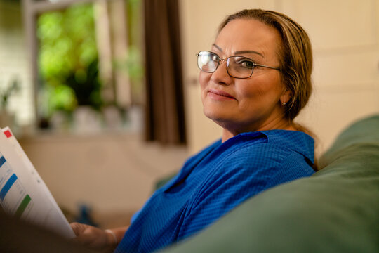 Woman sitting calmly on a couch while reading documents indoors during daytime, surrounded by natural light and greenery
