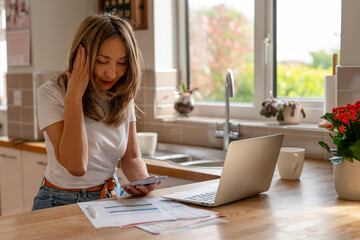 Woman experiencing stress while working on financial documents at home kitchen with laptop, phone, and plants in bright natural light during daytime