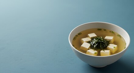 Minimalist composition of a warm bowl of Japanese miso soup with tofu and seaweed against a calm blue background with copy space