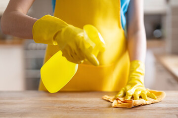 Person wearing yellow gloves sprays cleaner on a wooden surface while wiping it with a cloth in a home kitchen during daytime