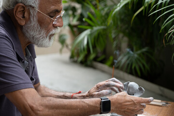 Indian old man crafting an eco-friendly clay Ganesh idol for Ganesh Chaturthi and teaching kids how to do it, sitting outdoors in a garden
