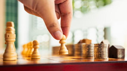 Hand moving chess piece with coins and wooden houses in background. Concept of financial strategy,...