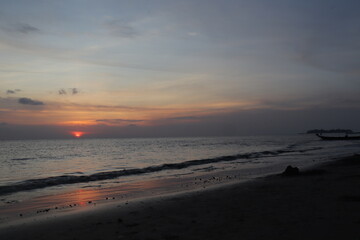 Sunset over calm ocean casting warm glow across the water. Beach sandy under sky is lightly clouded with hues of orange and pink blending into blue sea. 