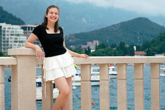 A young beautiful girl poses in summer on the resort's city promenade against the backdrop of the sea and mountains, dressed in a black top and a white short skirt - Powered by Adobe