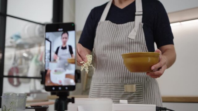 Asian woman mixes ingredients while streaming cooking vlog at home kitchen, smiling as she shares healthy homemade recipes with her audience.