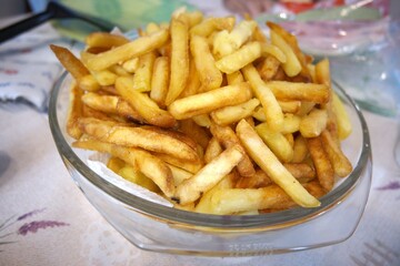 Golden crispy French fries served in a large transparent glass bowl on a table, close-up view.