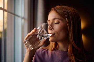 Woman with eyes closed drinking water by window.
