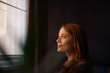 Fototapeta premium Smiling woman day dreaming by window at home.