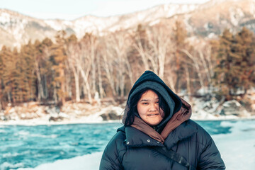 portrait of young Asian woman in black winter jacket with hood smiling outdoors against snowy mountain forest and blue river, concept of winter tourism, cold weather clothing, plus size