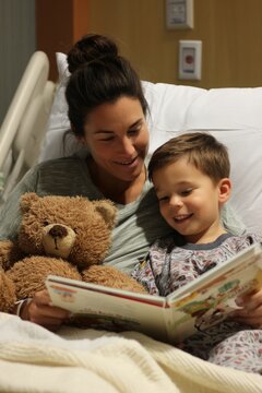 A mother reading to her child in a hospital bed. The child smiles while holding a stuffed animal. The mood feels comforting and hopeful, Generative AI